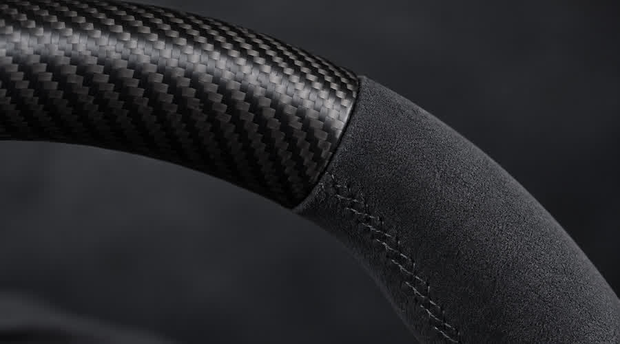 Macro view of a Porsche-style custom steering wheel showing the transition between satin-matte carbon fiber and charcoal microfiber grip material.